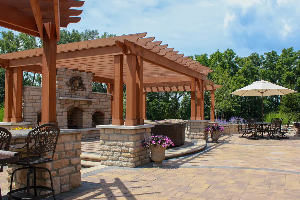 Pergola-framed fireplace patio with stone columns, paver surface, bar seating, dining space, and umbrella.